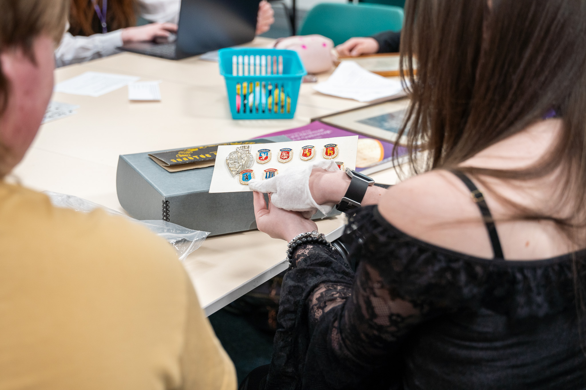 A student in gloves handles a card of mounted badges from an archive box, Prior Pursglove College.