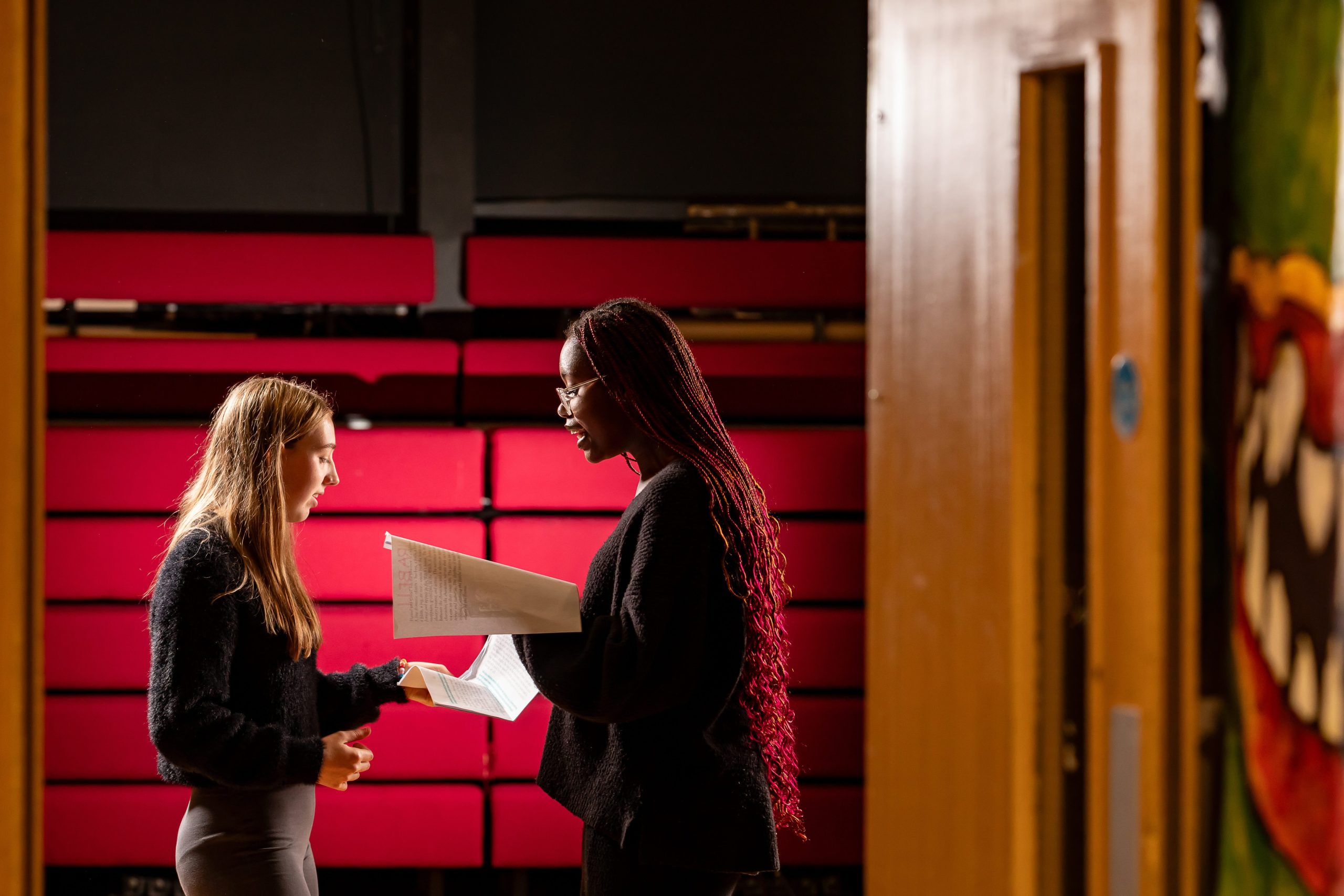 Two students standing in a theatre reading from a script, with red tiered seating behind them.