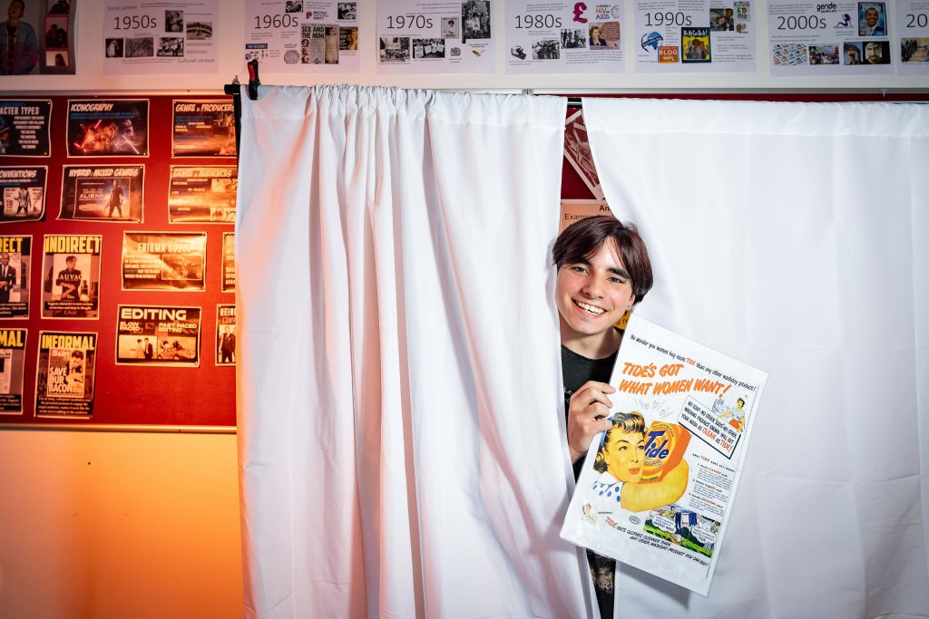 A student smiling from behind a white curtain and holding a printed vintage washing powder advertisement.