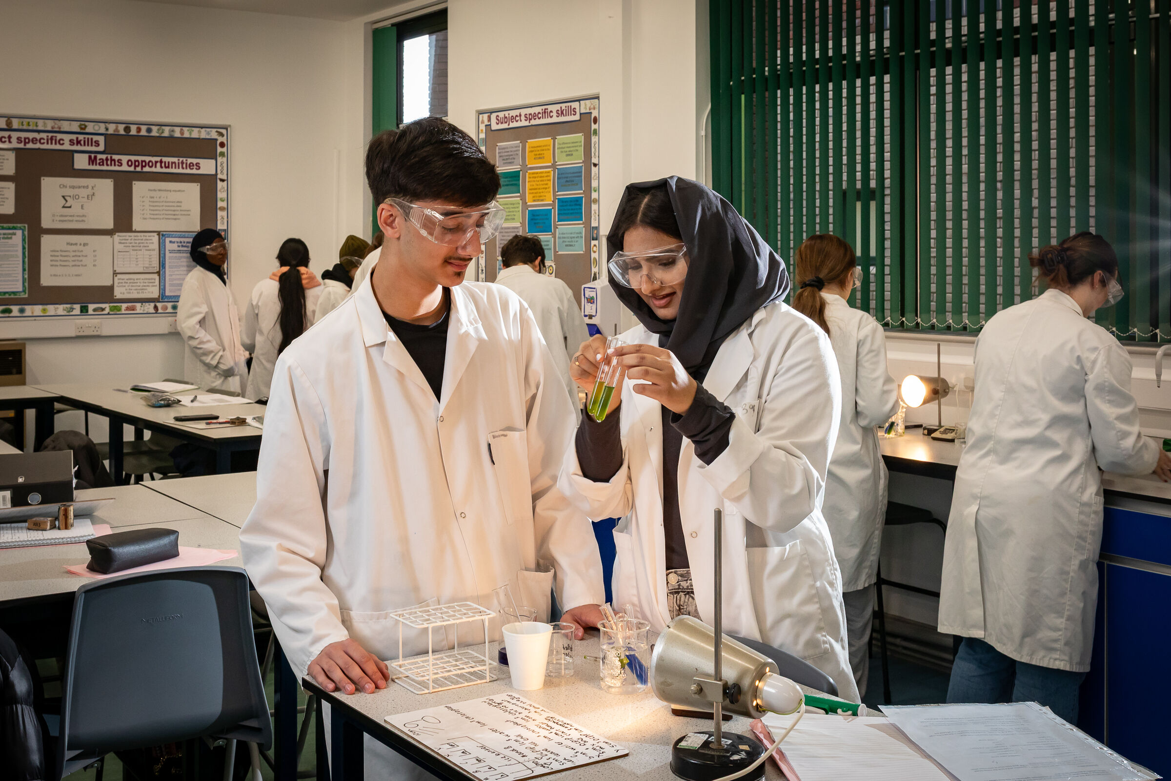 Two chemistry students in lab coats and safety glasses carrying out a practical at Prior Pursglove College