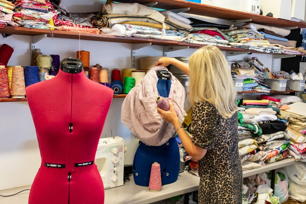 A woman in a patterned dress draping pale fabric over a tailor's dummy in a textiles room lined with shelves of material.