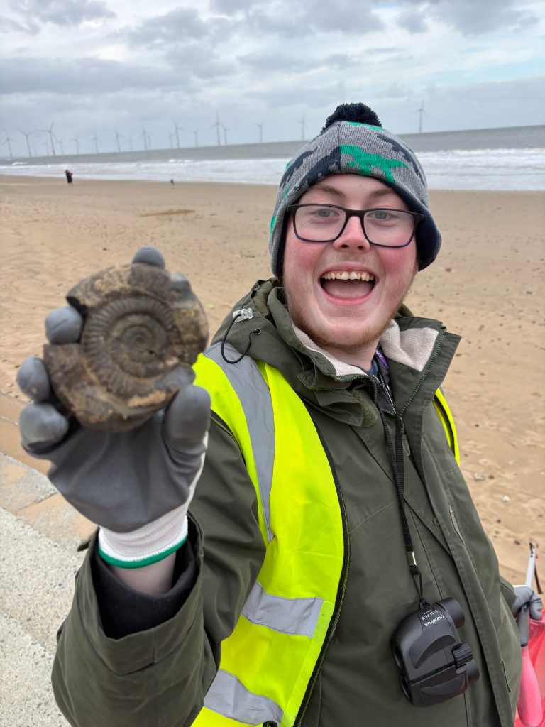 A smiling student in a high-visibility vest holds up a fossil found on Redcar beach