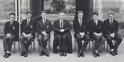 A master in a gown seated with six prefects in blazers in front of the Waterhouse arch, 1963.