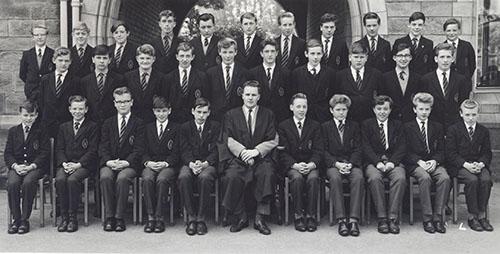 A class of students in blazers in three rows with a master in a gown seated in the centre, in front of the Waterhouse arch, 1963.