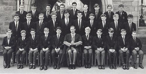 A class of students in blazers in three rows with a master in a gown seated in the centre, in front of the Waterhouse arch, 1963.