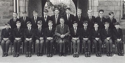 A class of students in blazers in two rows with a master seated in the centre, in front of the Waterhouse arch, 1963.