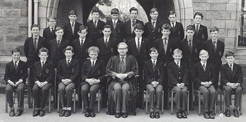 A class of younger boys in blazers in three rows with a master in a gown seated in the centre, in front of the Waterhouse arch, 1963.