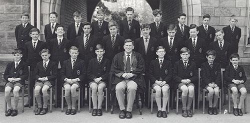 A class of younger boys in blazers in three rows with a master seated in the centre, in front of the Waterhouse arch, 1963.