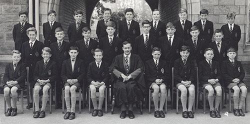 A class of younger boys in blazers in three rows with a master in a gown seated in the centre, in front of the Waterhouse arch, 1963.