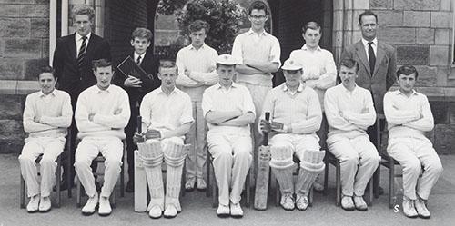 The 1963 cricket team in whites with bats and pads, in two rows in front of the Waterhouse arch, with two masters.